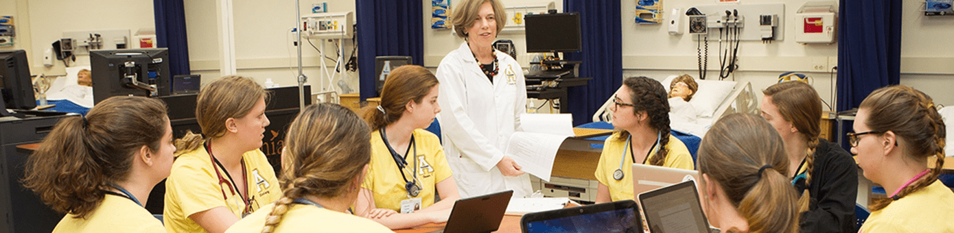 A medical training session with a group of students in yellow scrubs seated around a table with laptops, listening to an instructor in a white lab coat. Medical equipment and hospital beds are visible in the background, indicating a clinical learning environment.