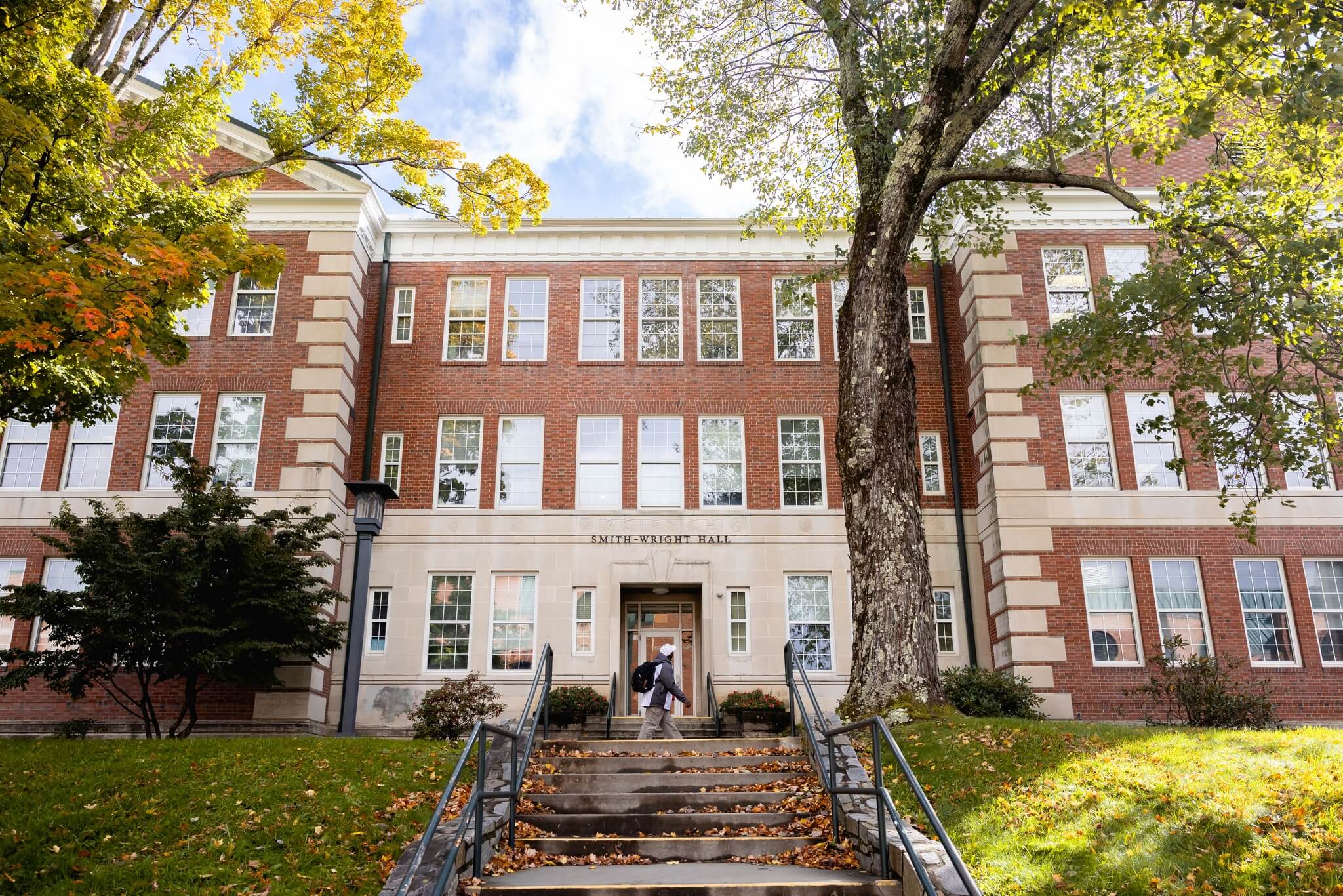 A student with a backpack walking by a three-story brick academic building surrounded by trees with autumn foliage.