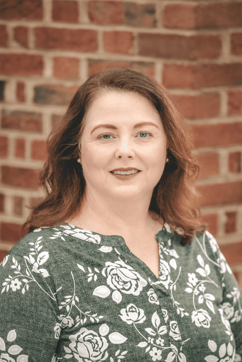 A headshot of a woman wearing a green and white floral top, with a brick wall in the background.